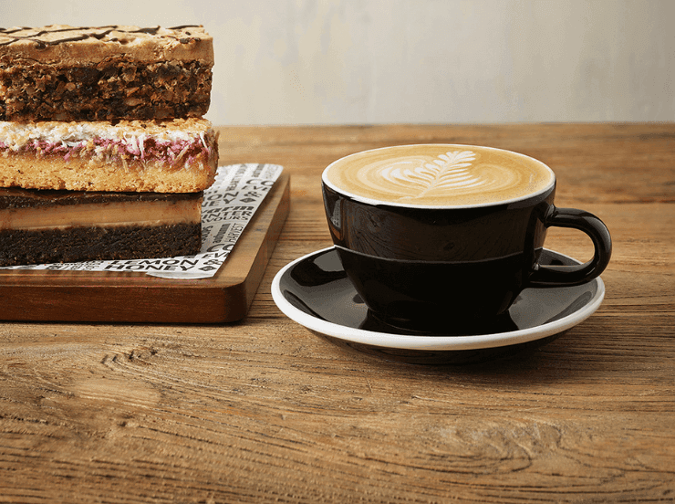 A black coffee cup with a flat white inside and three slices stacked beside on a board